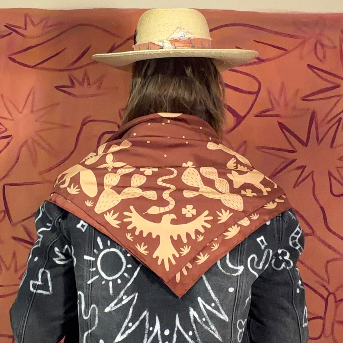A person viewed from behind wearing a tan cowboy hat, a black denim jacket with white hand-drawn style graphics, and a brown bandana draped over their shoulders, standing in front of a large orange artwork on a wall.