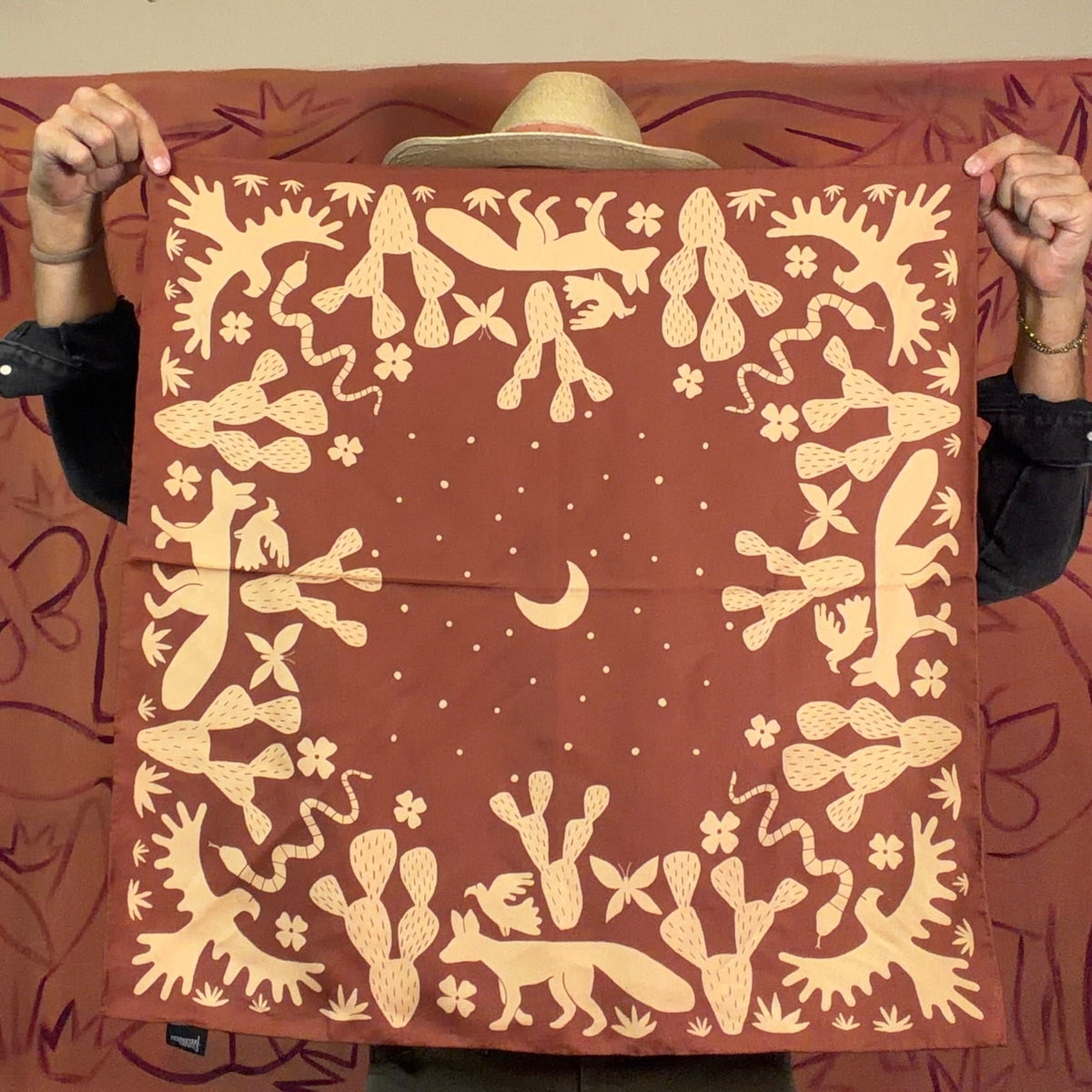 A person holds up a square scarf or bandana with a terracotta red background and a cream-colored abstract animal and floral pattern. The person is wearing a straw hat, a black long-sleeve shirt, and olive green pants, standing in front of a matching terracotta red fabric backdrop.