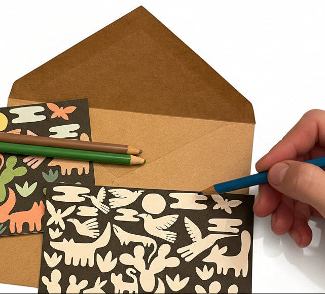 Hand holding a pen with two greeting cards featuring animal patterns and a brown envelope on a white background.
